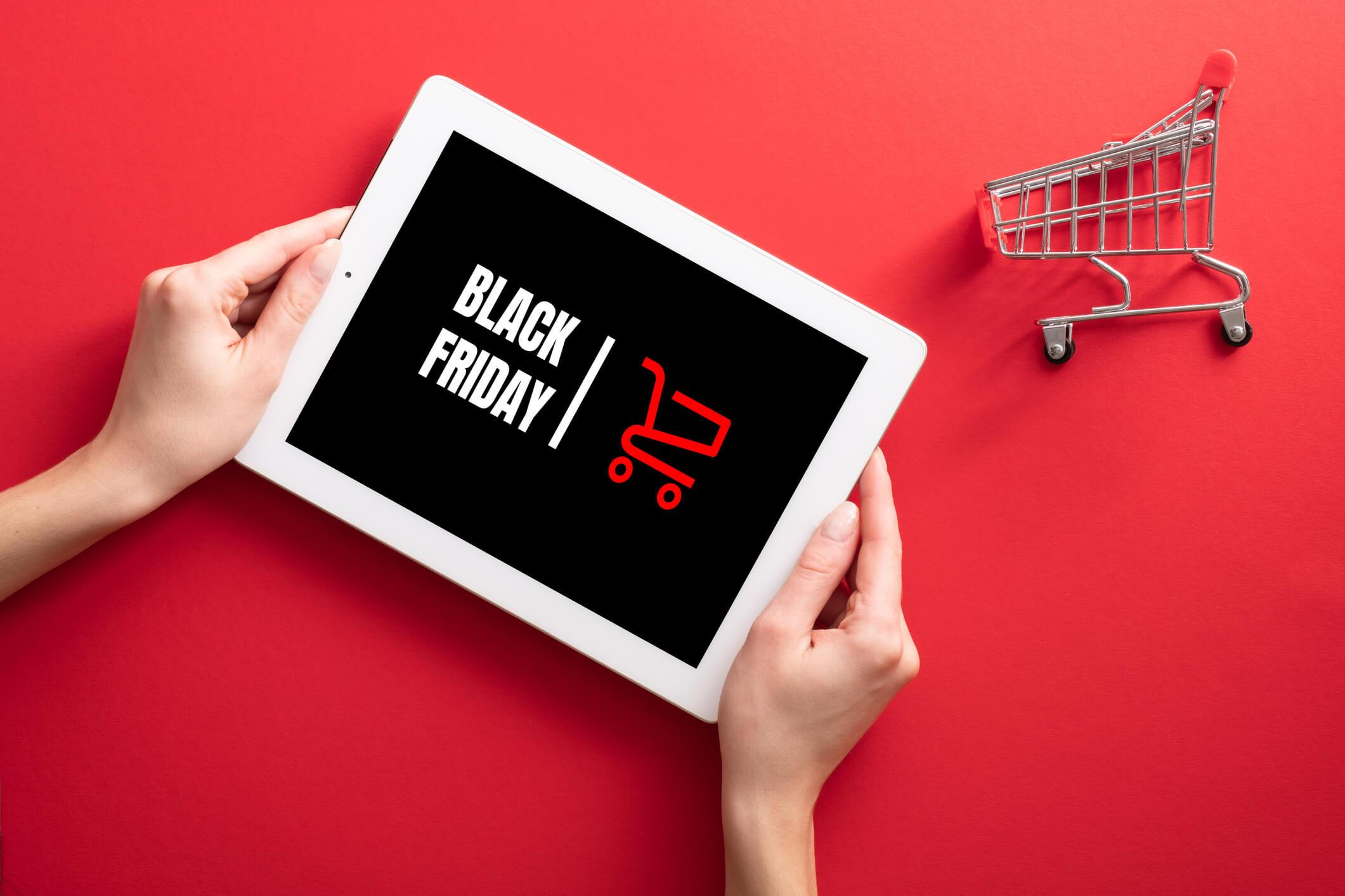  Close-Up Of Hand Using a Digital Tablet With Text that says Black Friday next to a small Shopping Cart Against Red Background 