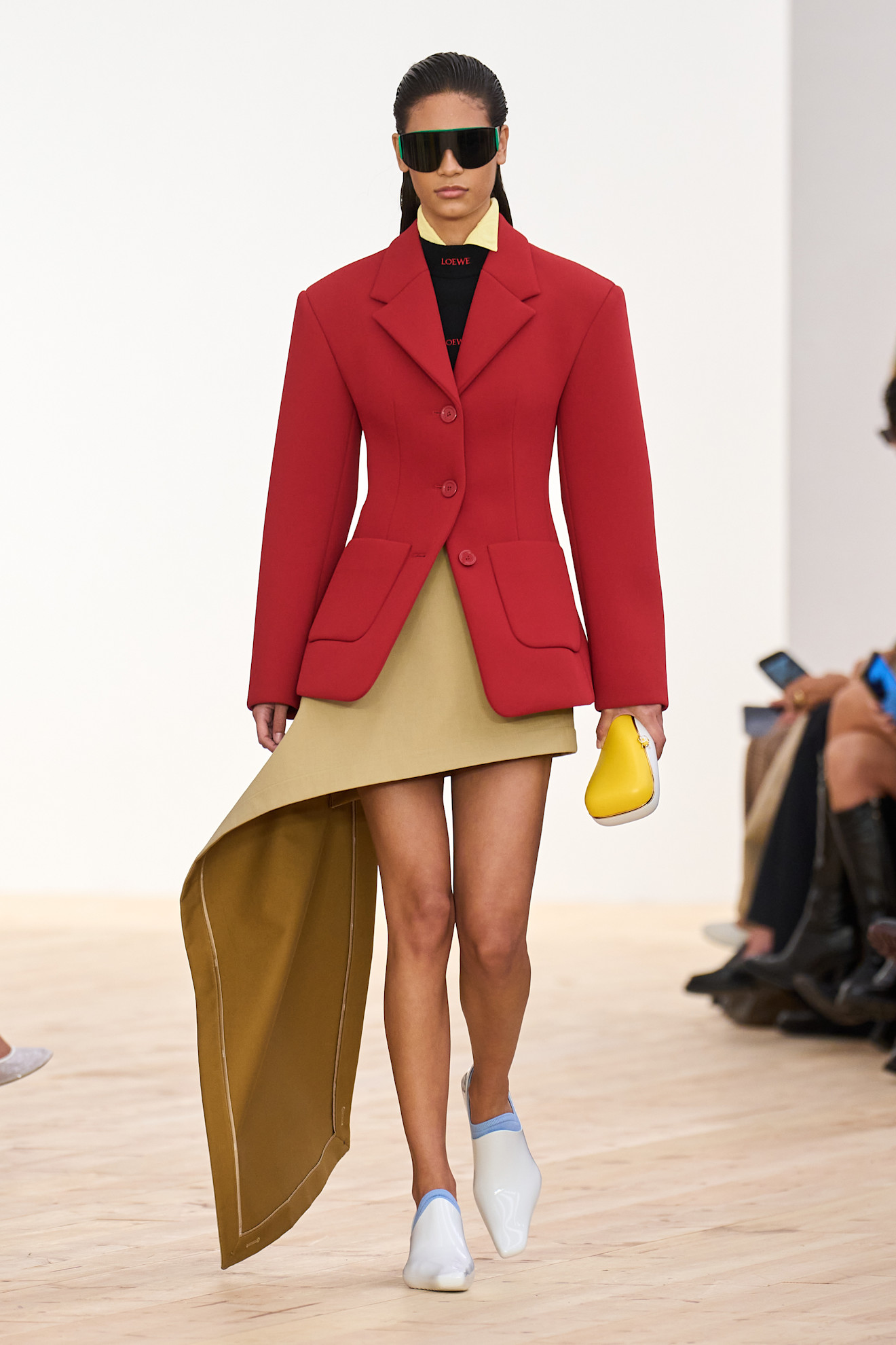 A Loewe model wearing a bright red blazer layered over a black logo crewneck shirt, a yellow collared shirt peeking out underneath, a khaki miniskirt with a train, a yellow clutch bag, white pointed loafers, and oversize black sunglasses.