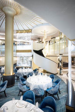Overview of Cunard’s Queen Anne Britannia Restaurant. Three round tables stand against a blue carpet and a golden column
