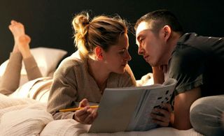 A woman (Kristen Stewart) lies on her stomach and holds a pencil and a magazine crossword puzzle, while a man (Steven Yeun) sits next to her and leans in near her face.