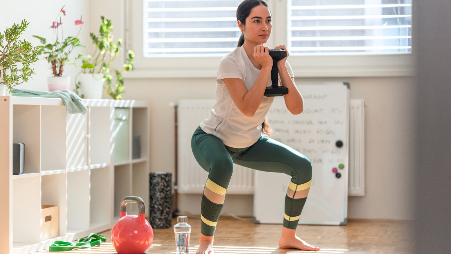 woman wearing green leggings and white tshirt performing a goblet squat in a home setting. there&#039;s a window and some white storage units behind her.