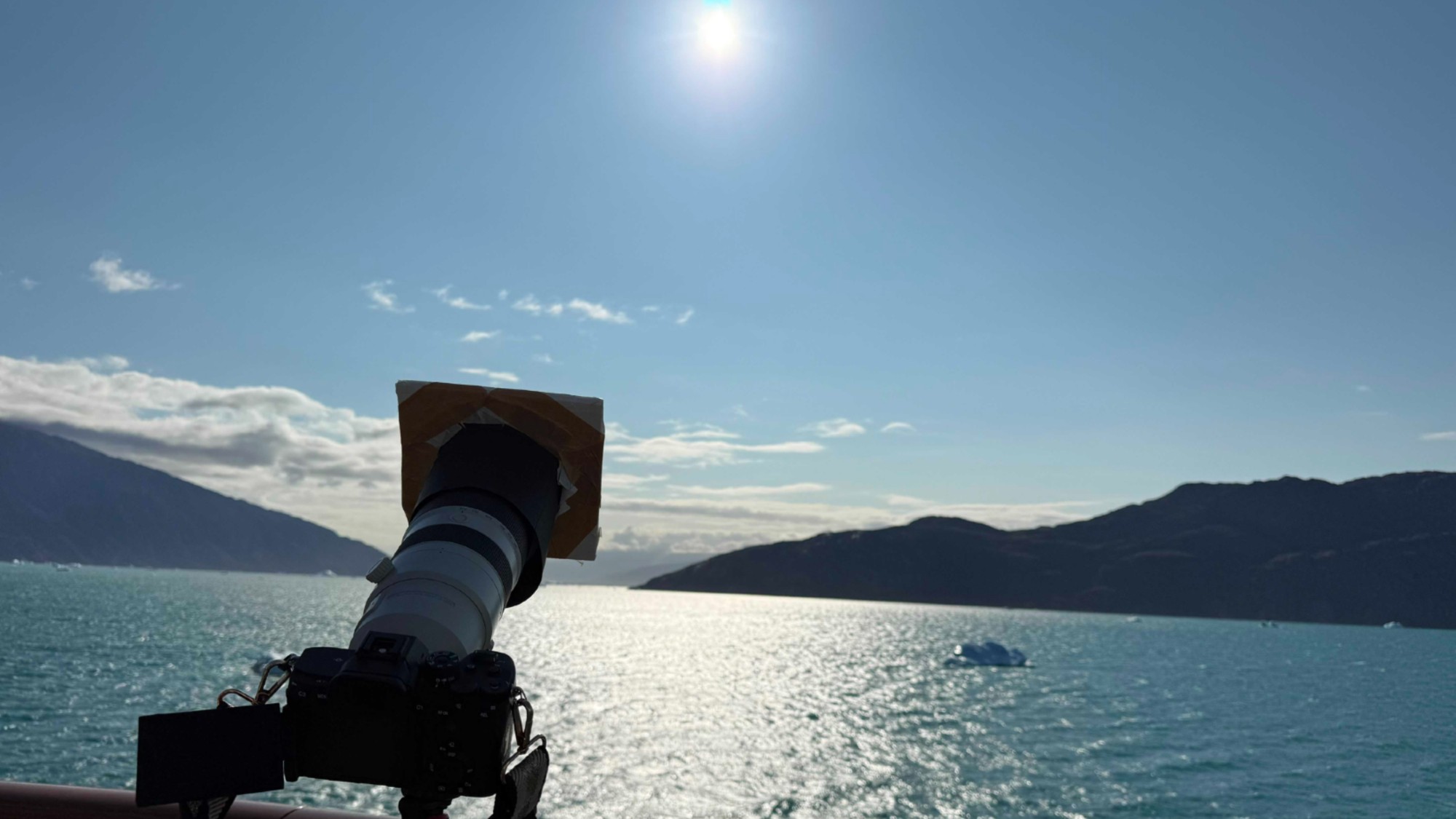 a camera pointed at the sun on the deck of a ship at sea. mountain peaks are visible in the distance