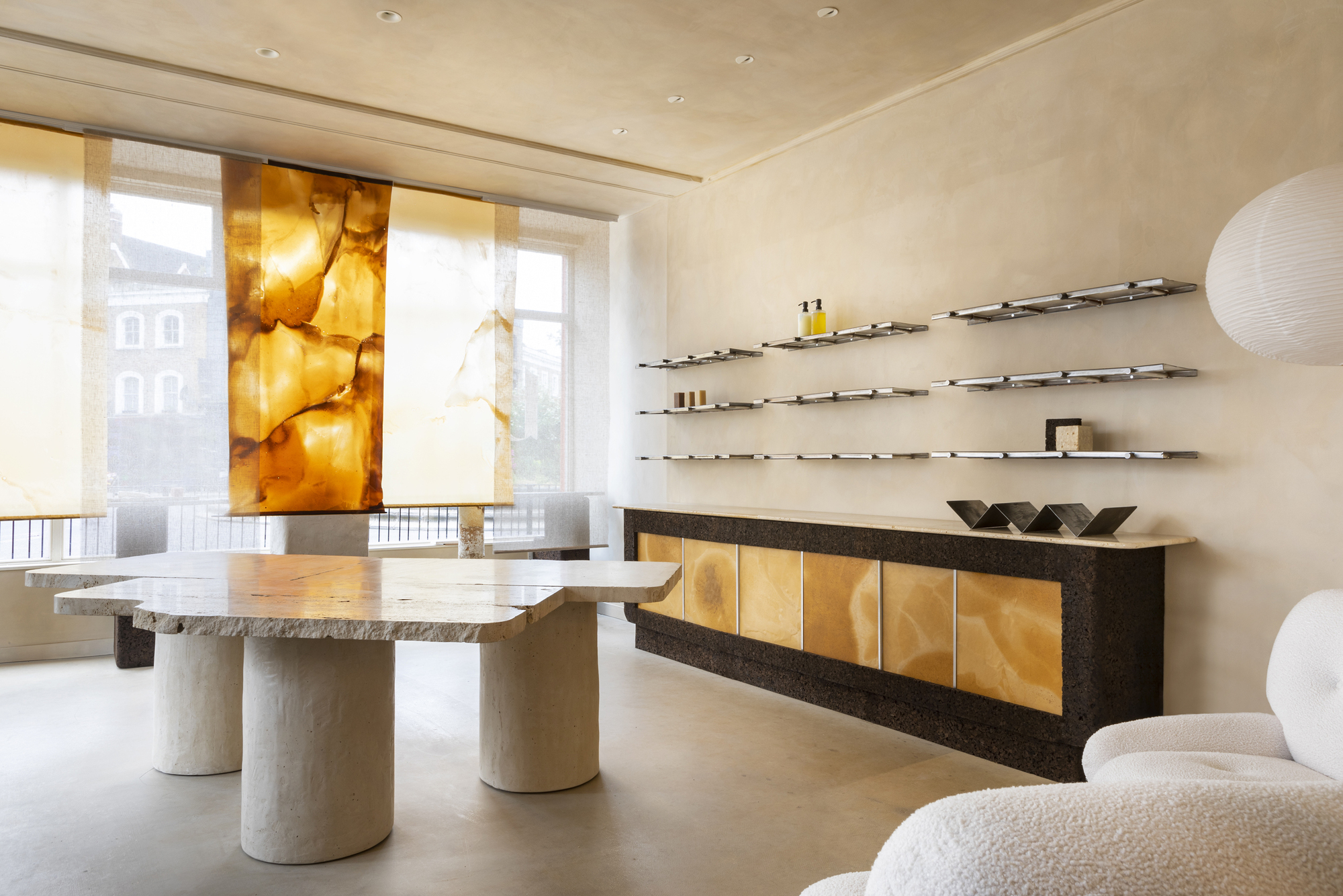 the interior of a beauty shop with limewashed walls, a travertine table, floating shelves over a long console table, and drapes hanging in the shopfront window