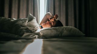 A woman lying on her back in bed, slowly waking up as a beam of light comes through a crack in the curtain