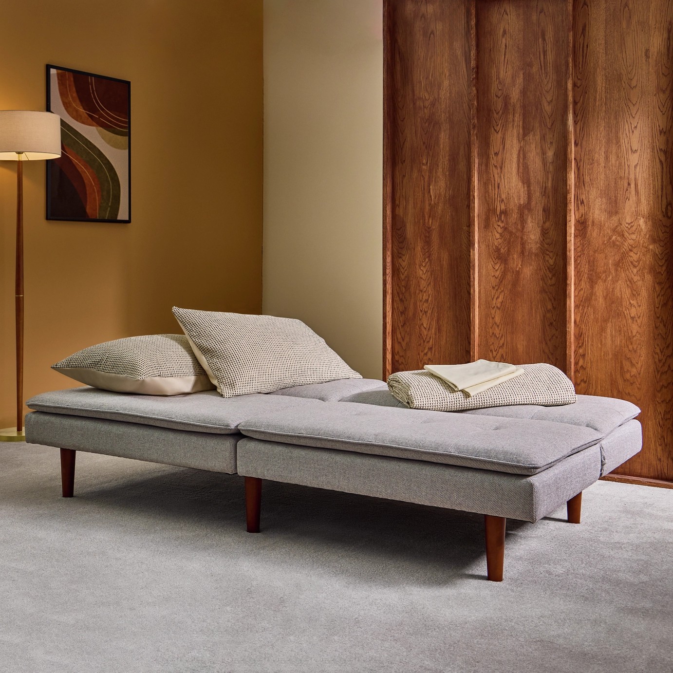 A gray, mid-century modern style sofa bed, extended as a bed in a room with light gray carpeting and walls that are half wood panelling, half yellow paint.