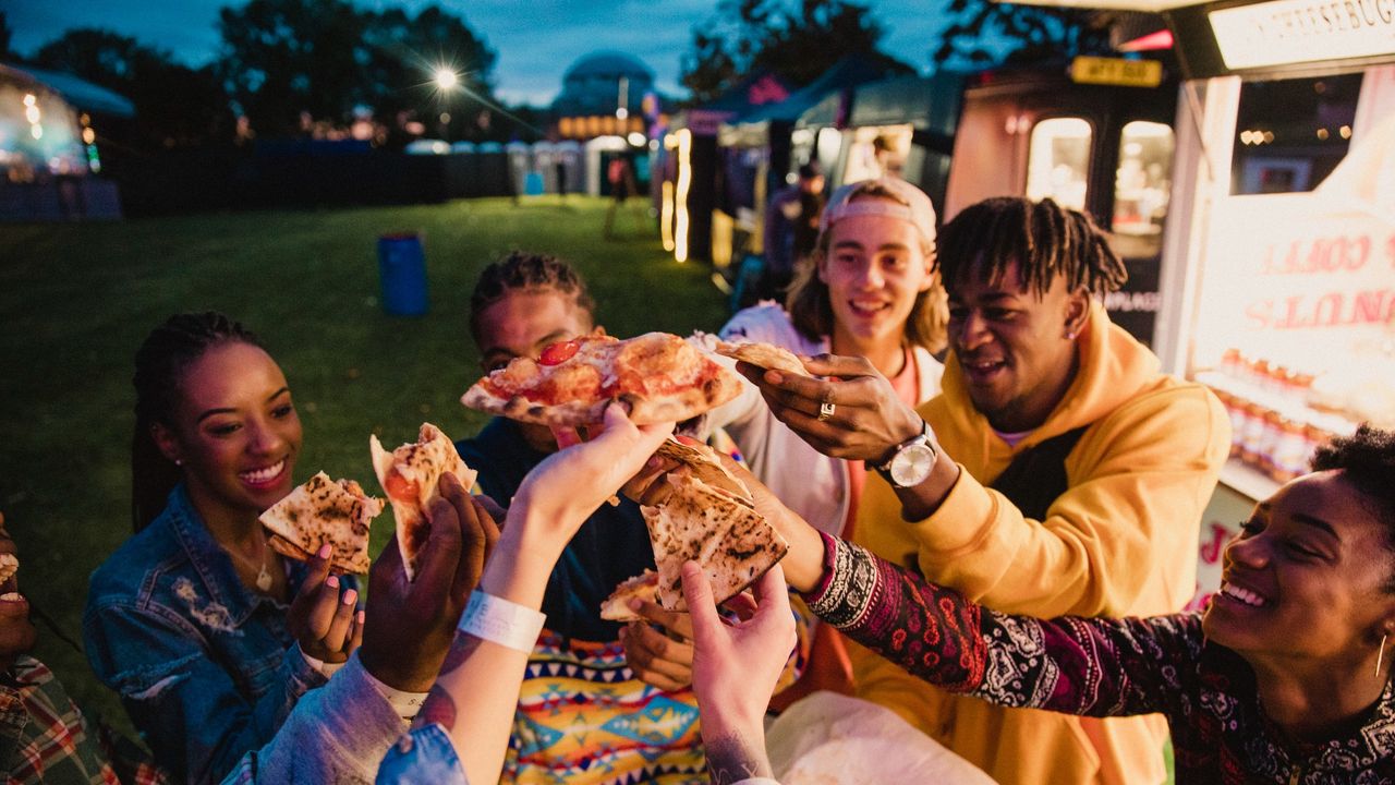 Group of young people sharing pizza in a field surrounded by food trucks