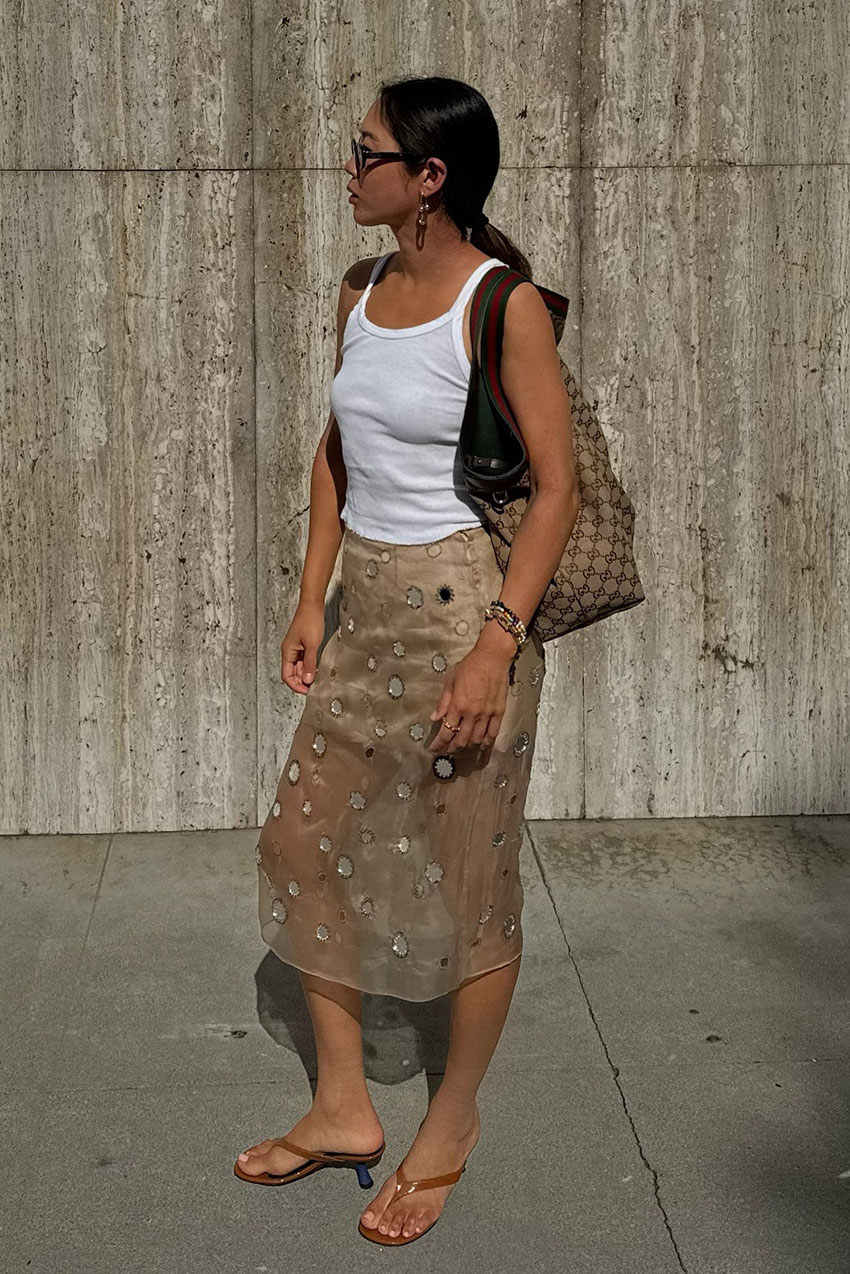 A woman wearing a white tank top, sheer Prada mirror skirt, and nude kitten heel thong sandals.