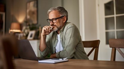 A man looks at his laptop while sitting at his dining room table.