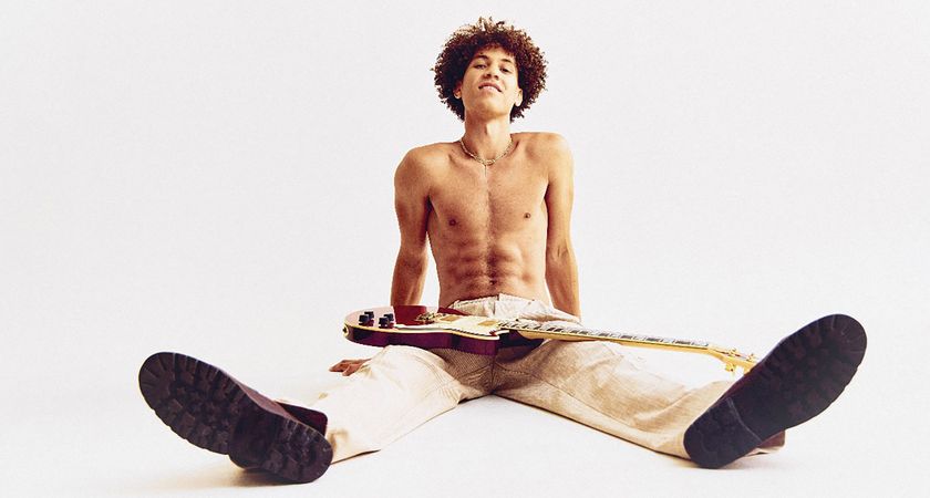 Brandon &#039;Taz&#039; Niederauer sits shirtless on a white floor, wearing ecru pants with a D&#039;Angelico electric guitar on his lap.