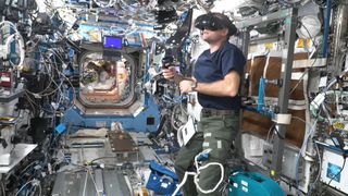 a person wearing a headset inside a very crowded international space station module with experiments covering every surface