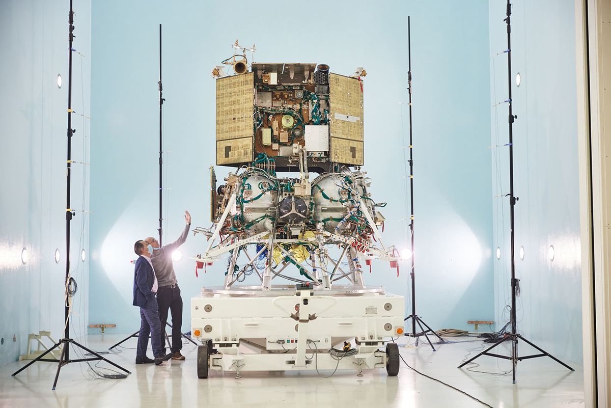 Two people stand next to Russia&#039;s Luna 25 moon lander in a test and assembly facility.