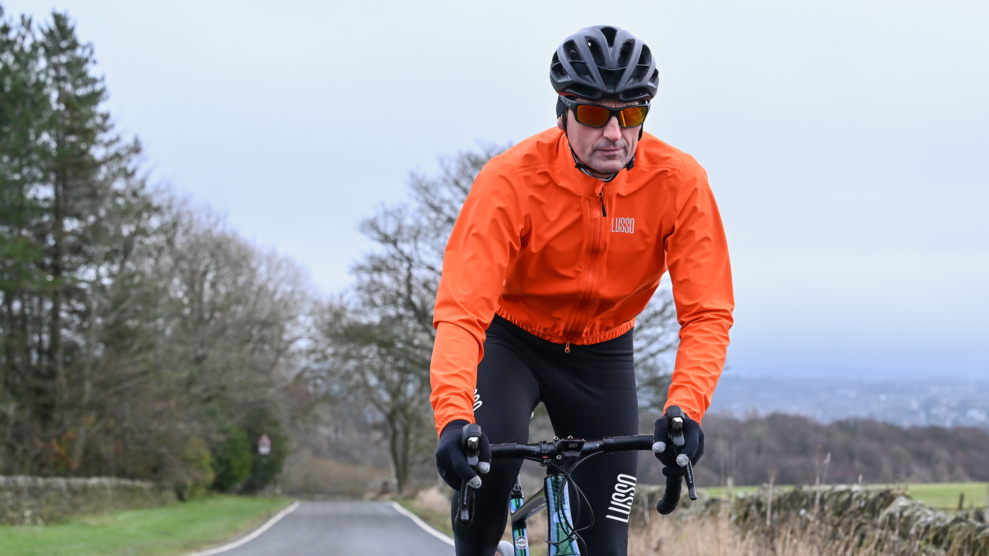 Man riding towards the camera on a green road bike wearing an orange jacket and black helmet.