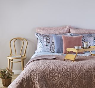 a pink bed and pillows in front of a white wall and a wooden chair next to it and a plant