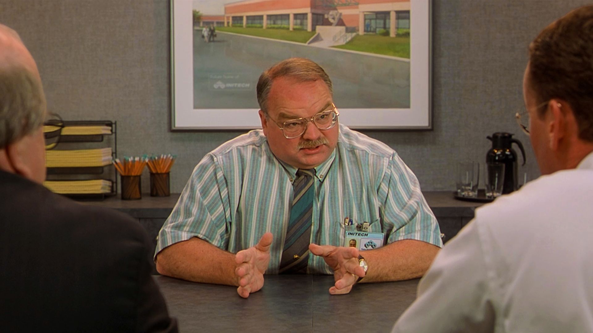 Riehle, dressed in a striped teal shirt and tie, wearing glasses, trying to explain what he 