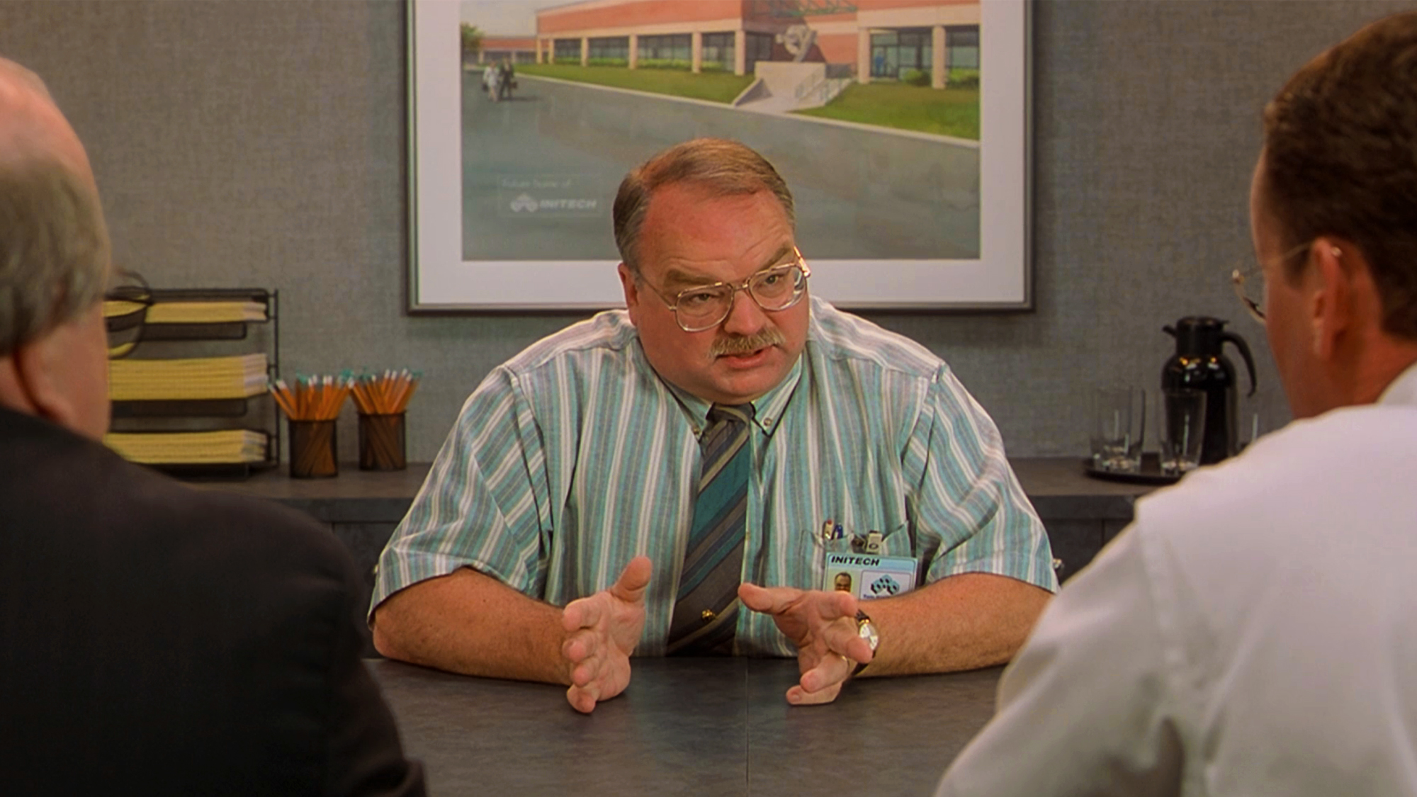 Riehle, dressed in a striped teal shirt and tie, wearing glasses, trying to explain what he "does here," in Office Space (1999).