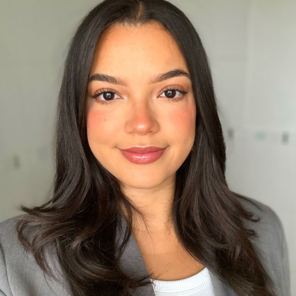 A woman with long dark hair smiles softly at the camera. She’s wearing a light grey blazer over a white top, with a blurred light background.