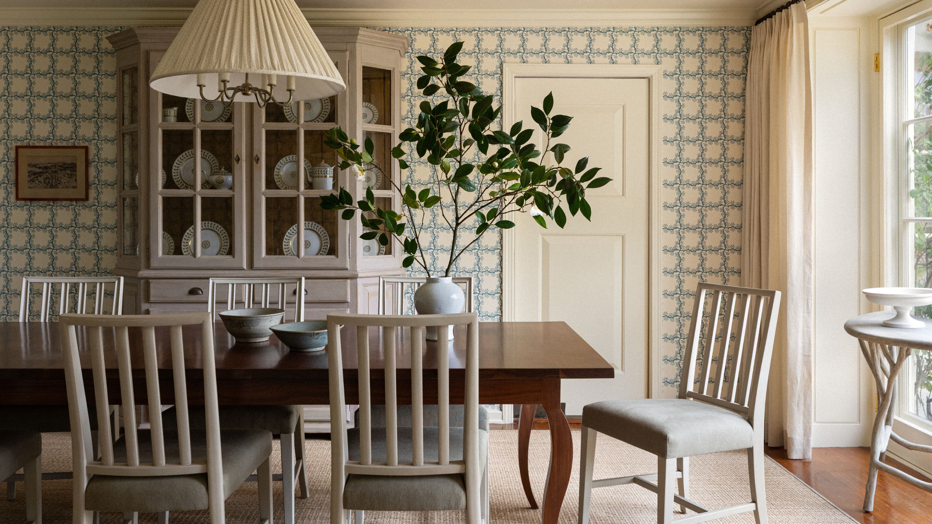 a wallpapered dining room with a soft cream and blue floral print, dark wooden table, white chairs and a vintage cabinet with china displayed in the background
