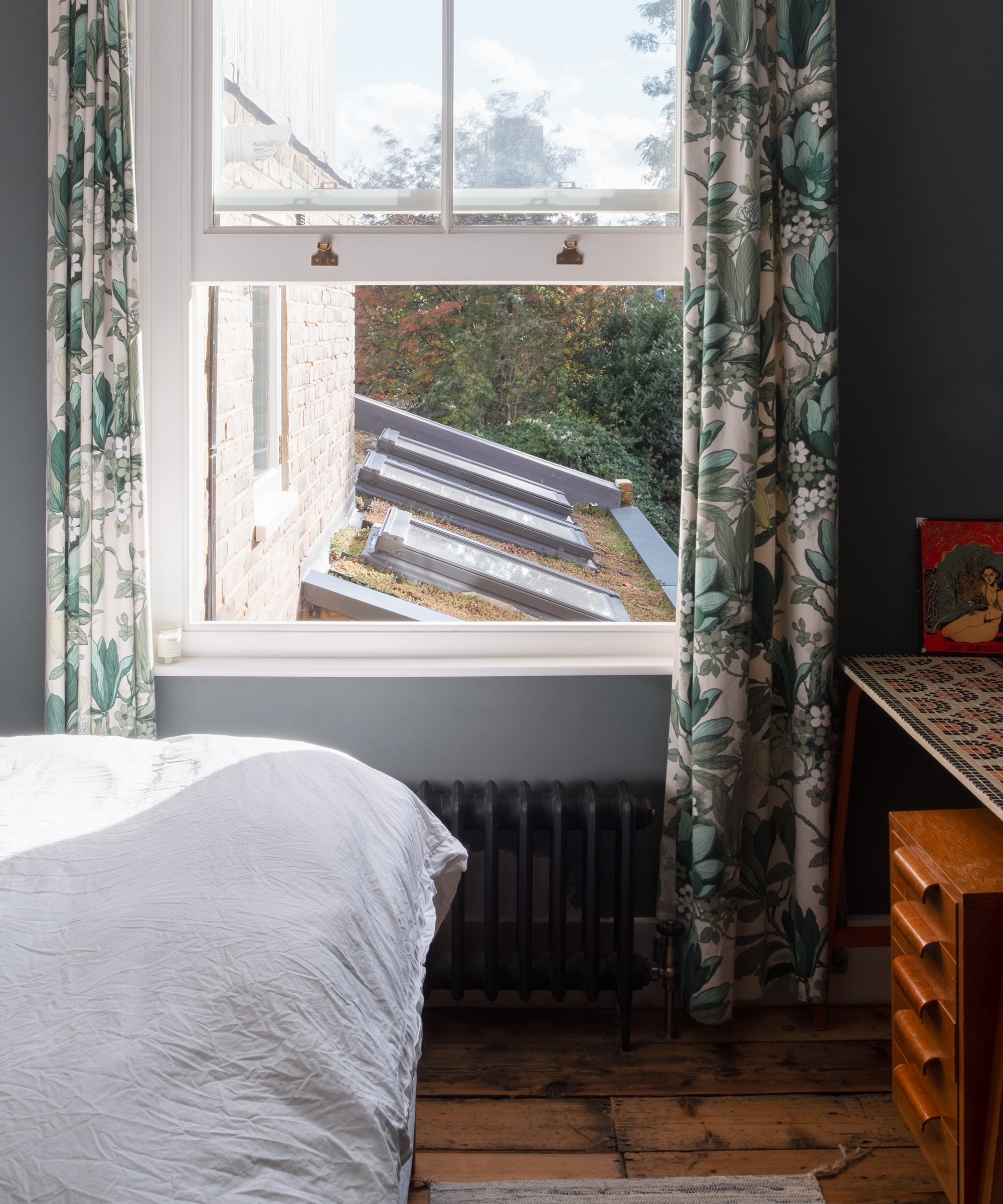 An open window in a bedroom with a green and white floral patterned print curtain window treatment. A retro-style desk is just visible to the right of the window and the base of a bed with a white cotton duvet cover