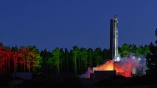 rocket on a launch pad with trees in behind. the trees are in artificial green and red colors from a nearby lighting source