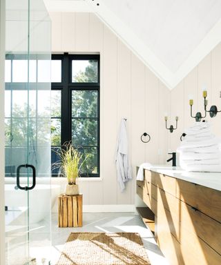 bathroom with neutral panelled walls, white sloped ceiling, black window frame, wooden vanity and corner of glass shower