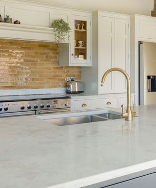 country-style kitchen with exposed wooden beams, cream cabinetry, and a large island with a stone worktop. Brass accents and a glazed tile splashback add warmth.