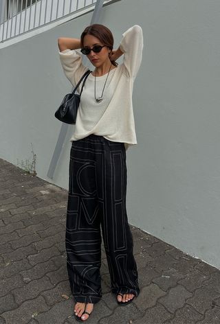 a photo of a summer rainy-day outfit with a woman wearing a white lightweight sweater over a tank with printed silk trousers, black strappy flat sandals, a cord necklace, black sunglasses, and a black shoulder bag