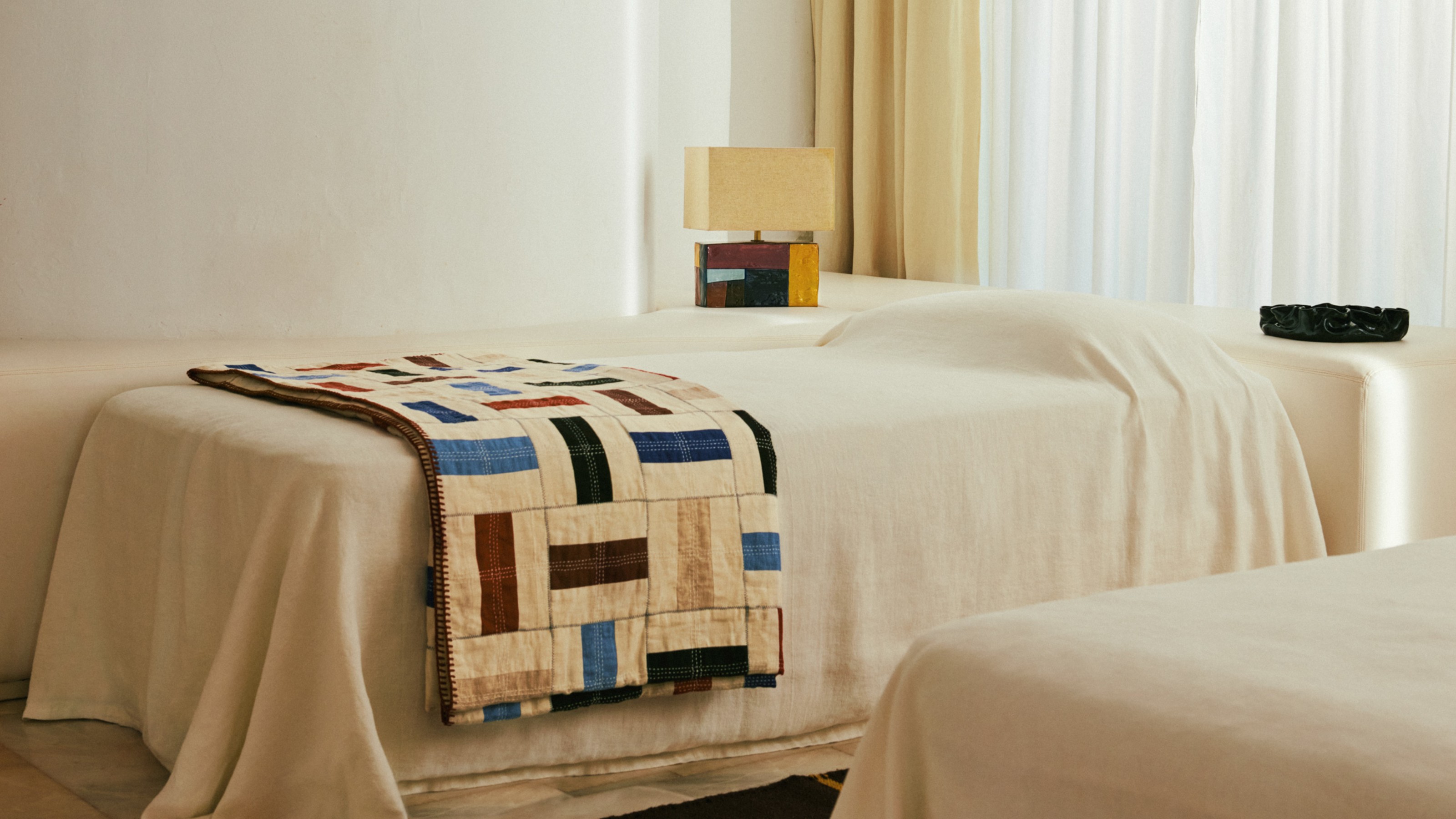 A minimalist bedroom with the bed covered in a white sheet with the Zara Home x Collagerie quilt on top and table lamp on the bedside table