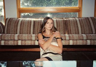 Actress Chloe East sits in front of couch wearing a cream dress with embroidered detailing.