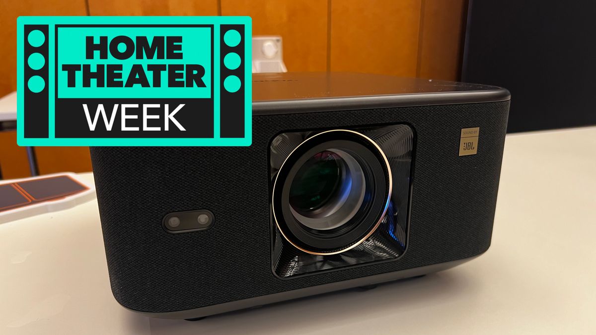 The Yaber K3 Pro projector showing the lens and front, on a white table, with a sign saying Home Theater Week