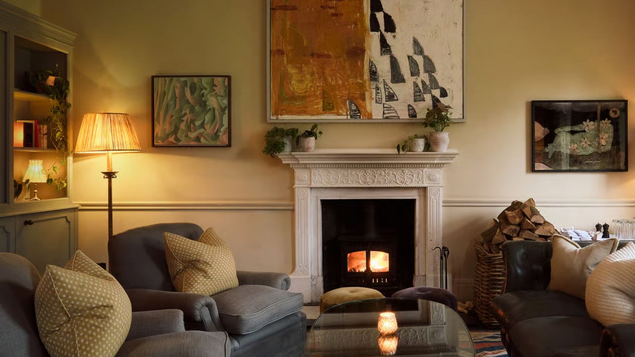 A living room with dim lighting with sofa and chairs in the space. There is also a fireplace with a large piece of artwork hanging above it. In the corner of the room is a floor lamp. 