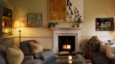 A living room with dim lighting with sofa and chairs in the space. There is also a fireplace with a large piece of artwork hanging above it. In the corner of the room is a floor lamp.