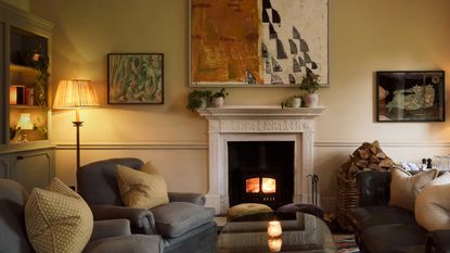 A living room with dim lighting with sofa and chairs in the space. There is also a fireplace with a large piece of artwork hanging above it. In the corner of the room is a floor lamp.