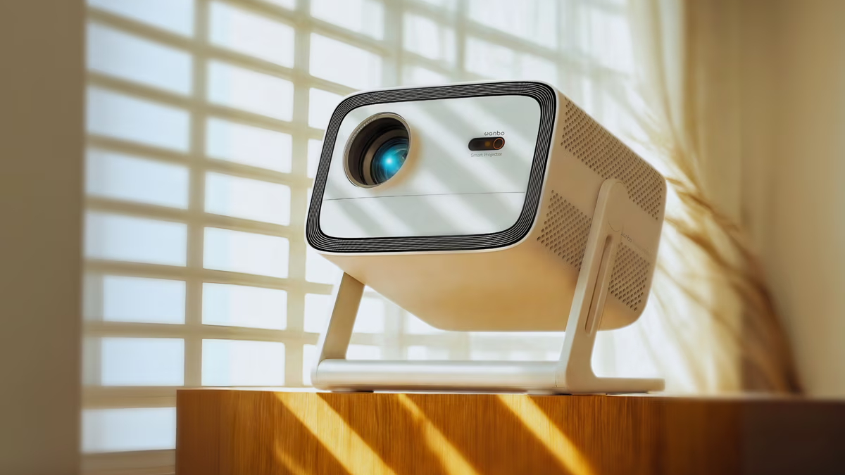 A white projector, the Vali 1, sits on a wooden tabletop. Behind the projector is the silhouette of blinds against the sunlight.