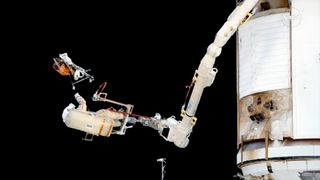 A spacesuited cosmonaut standing on a platform at the end of a robotic arm outside the International Space Station releases a bundle of trash into the blackness of space.