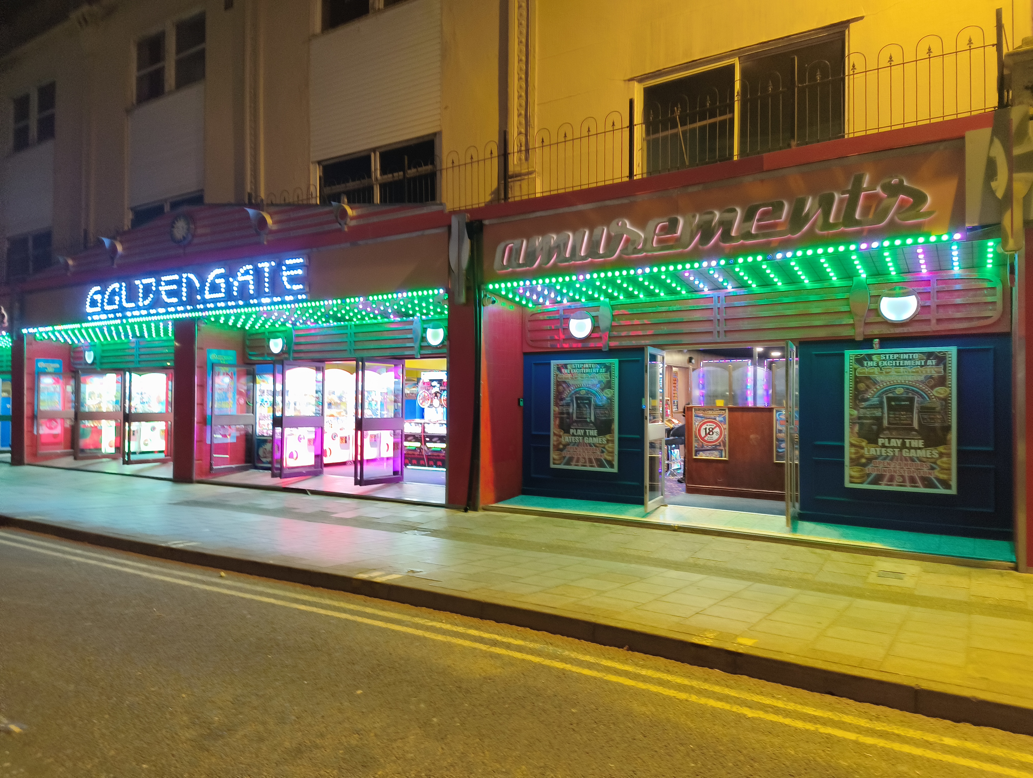 A nighttime view of two brightly lit, side-by-side amusement arcades, "Golden Gate" and "Amusements," with colorful neon and LED signs above the storefronts.