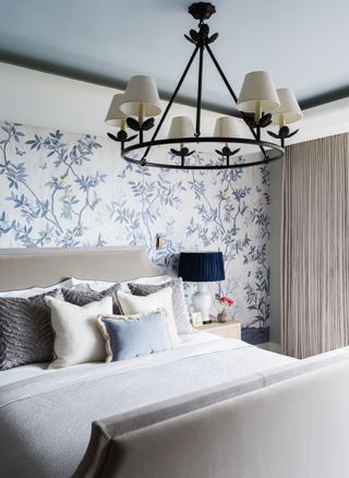 Bedroom by Elicyon with blue and white wallpaper, upholstered taupe bed, chandelier with shades