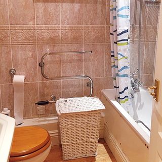 An outdated bathroom with neutral tiles and wood-effect toilet seat, shower in white bath with colourful patterned shower curtain decor