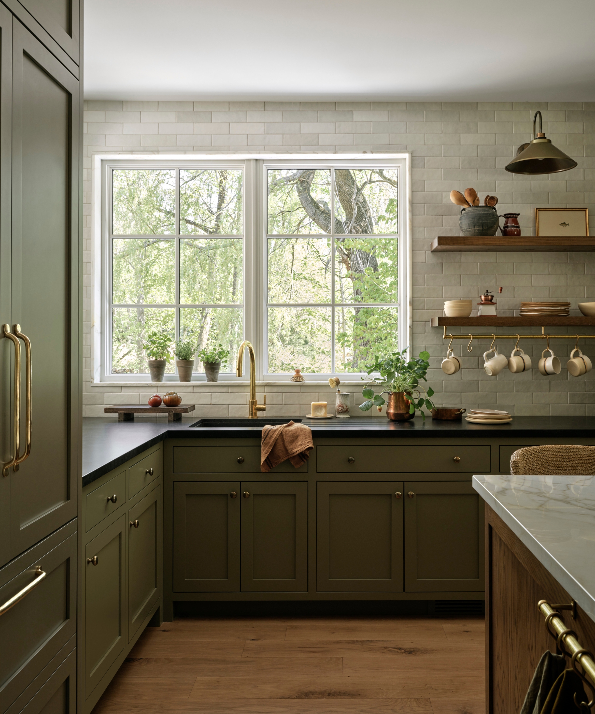 A kitchen with dark olive green cabinets, black countertops, brass hardware, and wooden floors