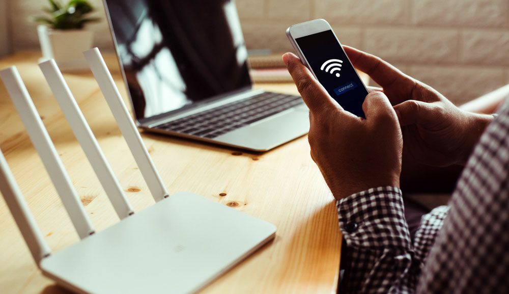 A man uses a smartphone to connect to a Wi-Fi network, with a router and a laptop sitting on a desk.