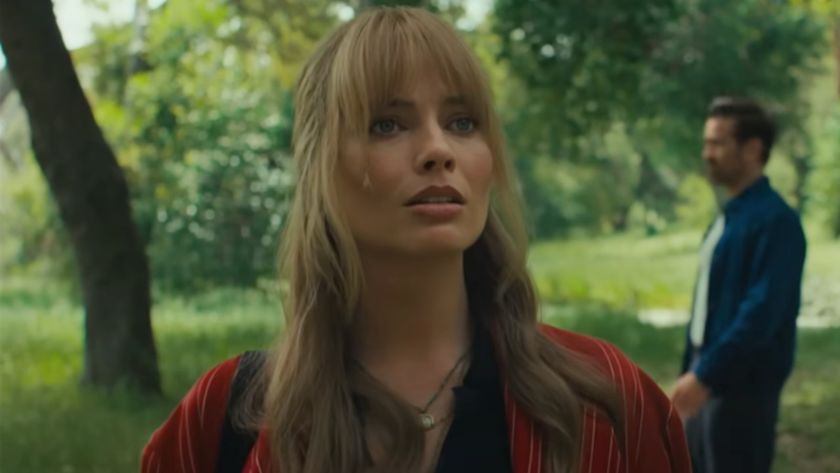 Margot Robbie looking curiously forward with Colin Farrell walking in the background behind her on the right. 