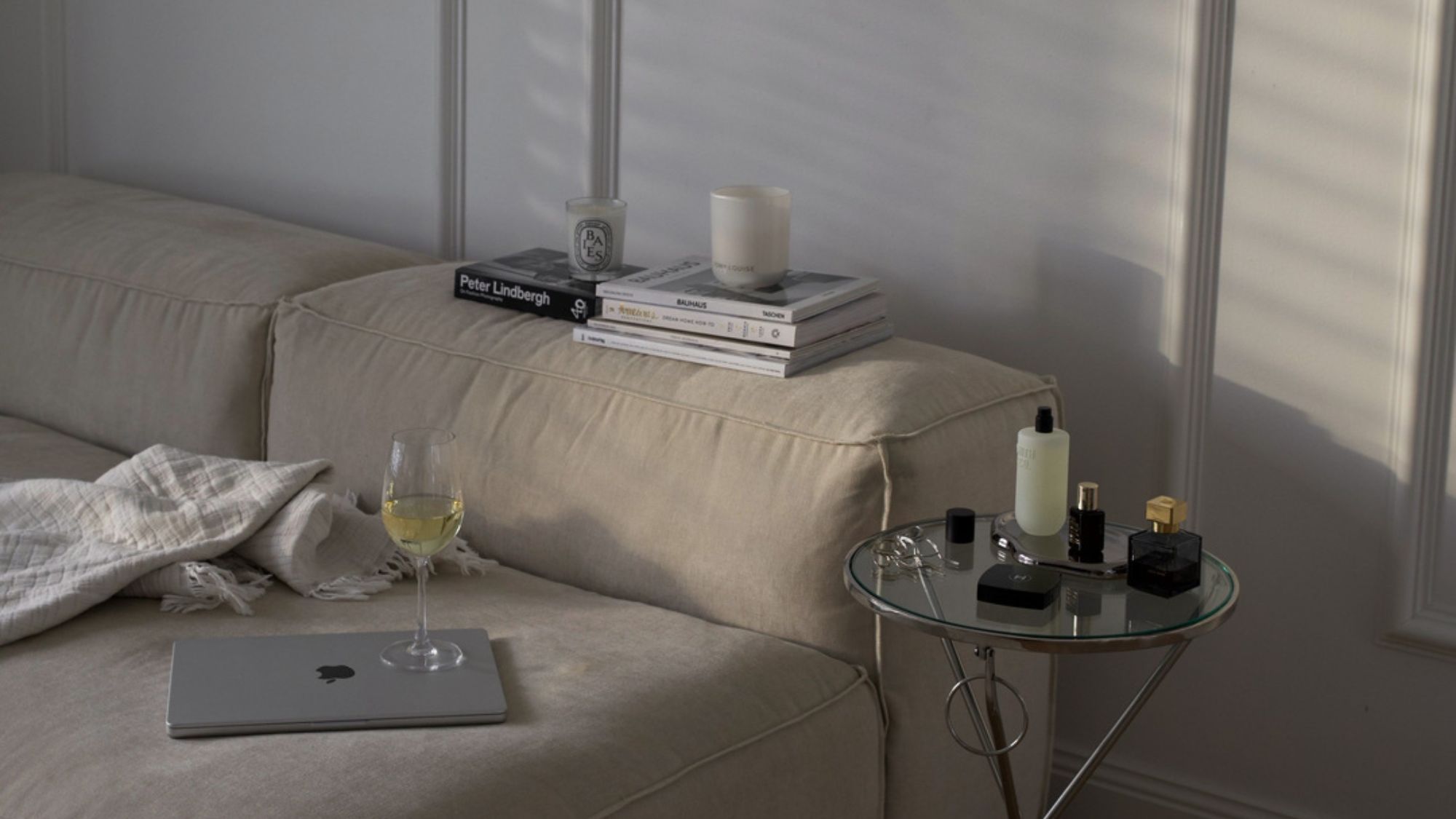 An image of a couch soaked in daylight with magazines, a laptop, and wine on top and beauty products on the side table.