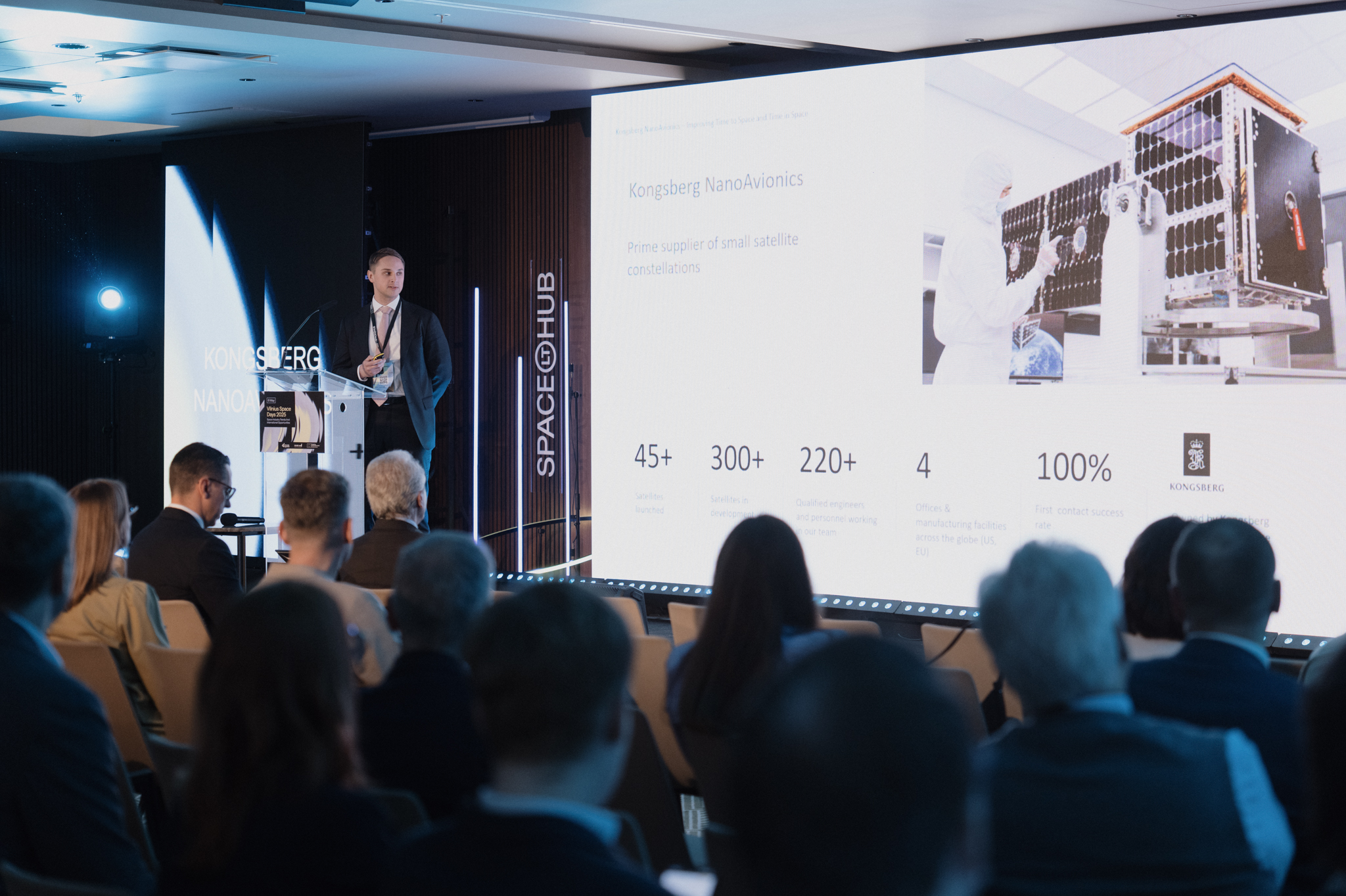 A man with a powerpoint in the background. An audience sits in front.