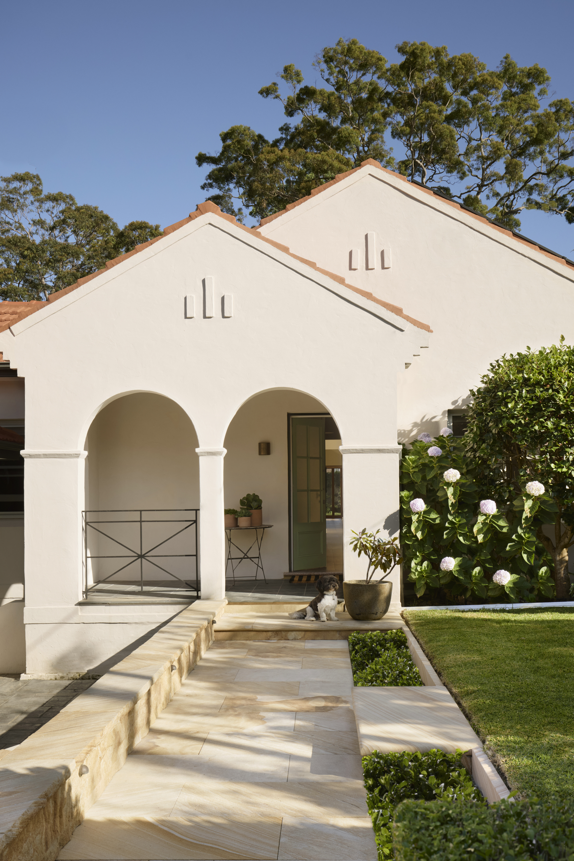 Image of the front of a white house that has two archways leading to a front stoop with a sage green door that is halfway open. There is a stone walkway and a grassy front yard