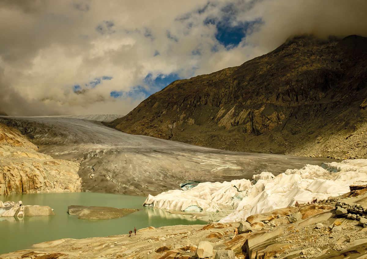 A grey, shrinking glacier flows down between rocky mountains into a milky green lake, with its lower edge draped in white protective blankets and tiny figures of visitors on the rocks
