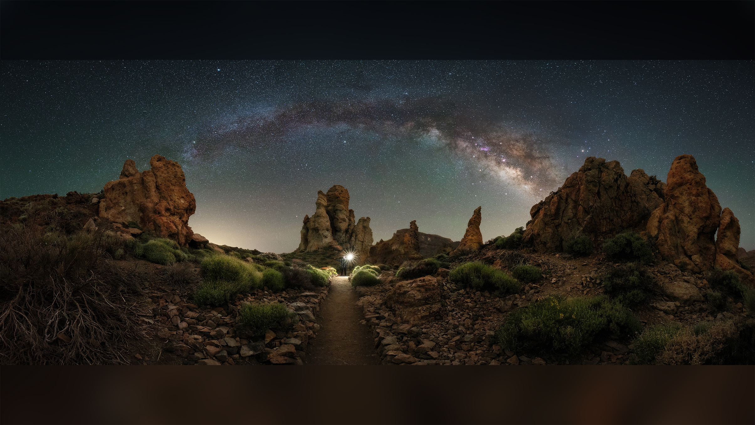 A stunning night sky panorama featuring the Milky Way over rugged rock formations and a winding path illuminated by a bright light