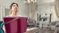 A woman holds a bucket up in her dining room to catch drips from a leaky roof.