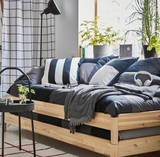 an Ikea sofa/day bed with wooden slats and lots of dark blue bedding and white and blue cushions, against a window, and a check white and black curtain in the corner