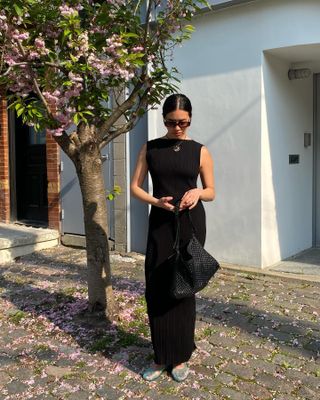 Sasha Mei wearing a black high-neck maxi dress with sunglasses, a braided bag, and blue jelly shoes.