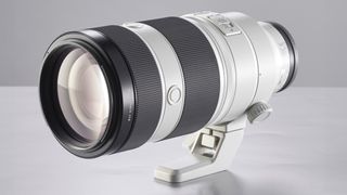 A sleek, silver-and-black camera lens rests on a light surface, featuring a tripod mount and textured grip sections for stability and control