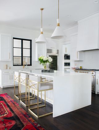 white kitchen cabinetry and island with dark wooden floor dressed with red and black turkish rug and statement white and gold pendant lights above island